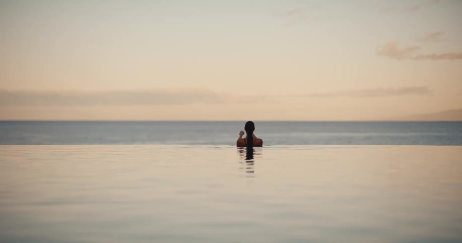 A woman peacefully ties her hair up while relaxing in a serene infinity pool overlooking the ocean at sunset