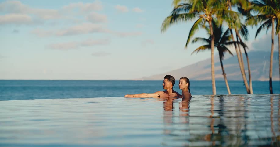A romantic couple relaxes together in a stunning infinity pool, overlooking the ocean under tropical palm trees
