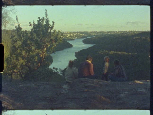 Vintage footage shows a family of four sitting on a rocky overlook, enjoying the scenic view of a winding river and a distant city skyline