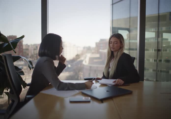 Two professional businesswomen collaborate during a meeting in a modern conference room with a large window overlooking the city