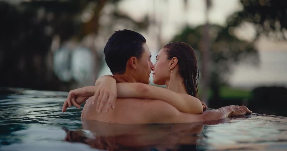 An attractive young couple embraces and kisses in a luxurious infinity pool at sunset