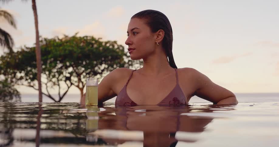 A serene woman in a bikini holds a glass of champagne while relaxing in an infinity pool overlooking the ocean at sunset