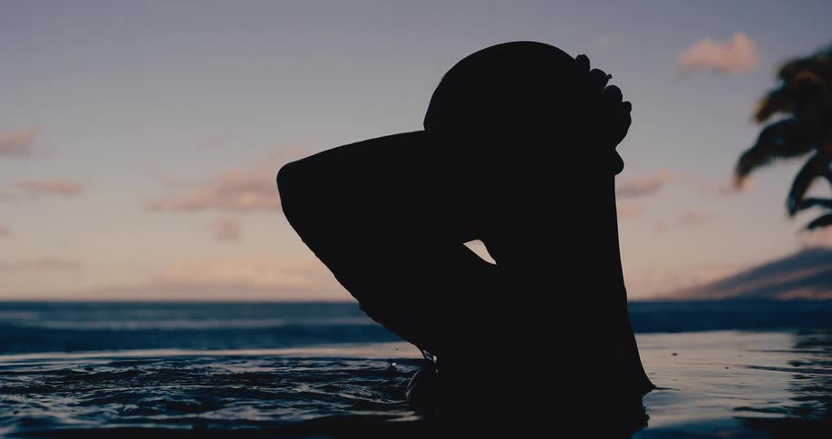 A woman's silhouette emerges in slow motion from an infinity pool at sunset, gracefully tying her wet hair with the ocean in the background