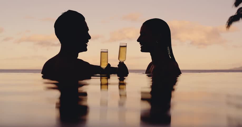 Silhouette of a romantic couple toasting with champagne glasses while relaxing in an infinity pool at sunset