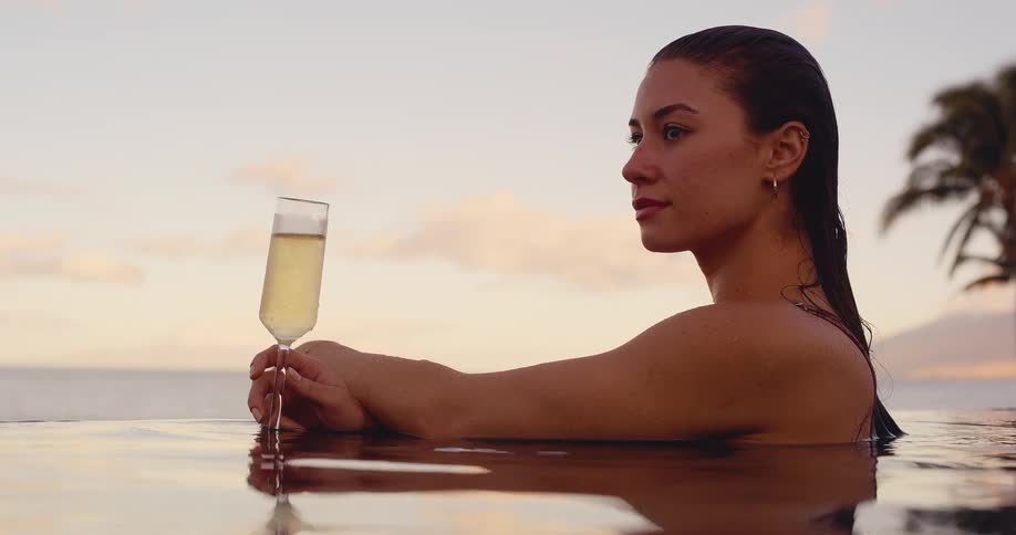 A woman relaxes with champagne in a tropical infinity pool, watching the sunset over the ocean