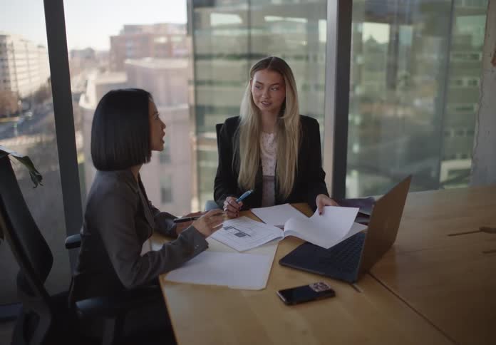 Two diverse businesswomen collaborate in a modern office, reviewing documents and using a laptop with a city view in the background