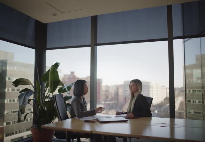 Two diverse businesswomen collaborate during a meeting in a modern, sunlit conference room with large windows overlooking the city