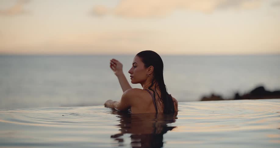 A beautiful woman with wet hair peacefully watches the sunset from a luxurious infinity pool overlooking the ocean