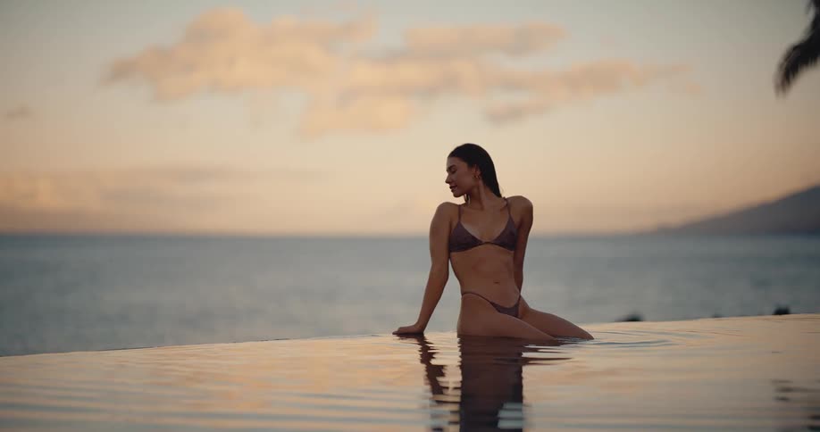 A beautiful woman in a bikini relaxes on the edge of an infinity pool, smiling and enjoying the tranquil sunset view over the ocean