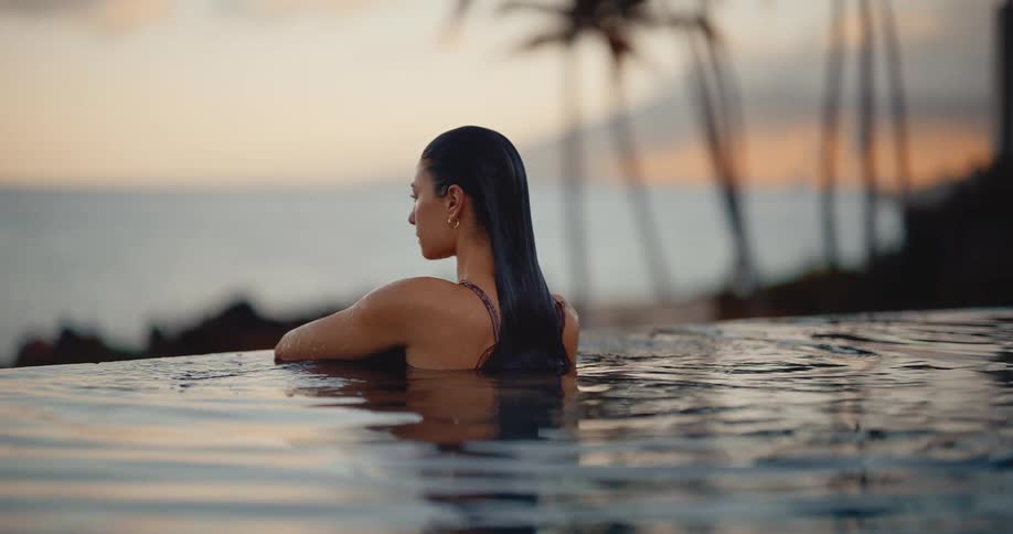 A serene woman rises slowly from the water in a tranquil infinity pool, relaxing at sunset with an ocean view