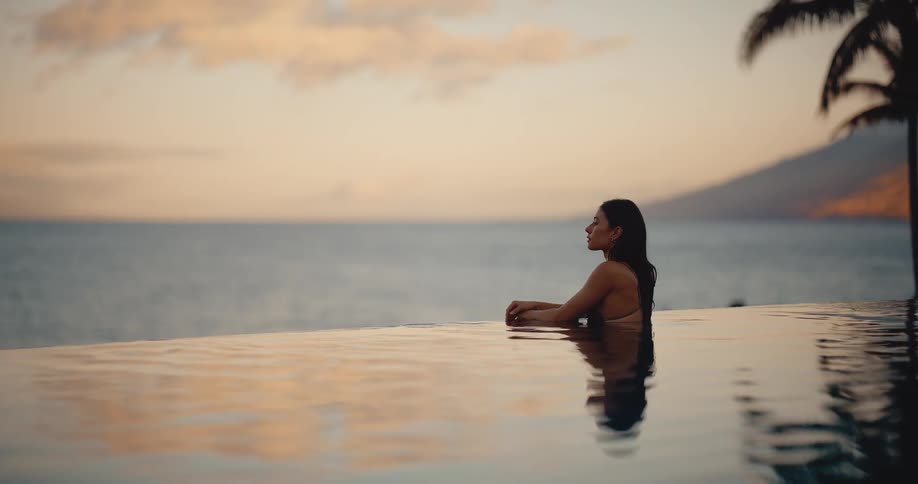 Woman peacefully relaxes in a resort infinity pool while watching the beautiful sunset over the ocean