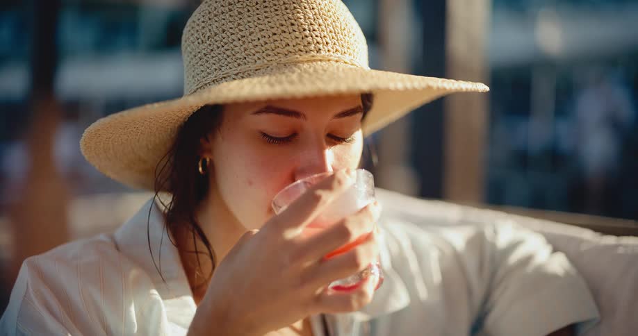 A stylish woman in a straw hat smiles and slowly sips a pink cocktail, enjoying the warm glow of the golden hour