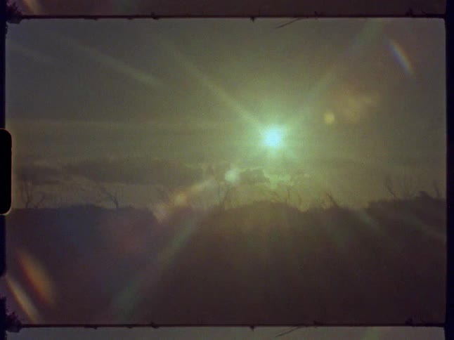 Old film footage shows a sun setting with a bright lens flare behind a silhouetted treeline