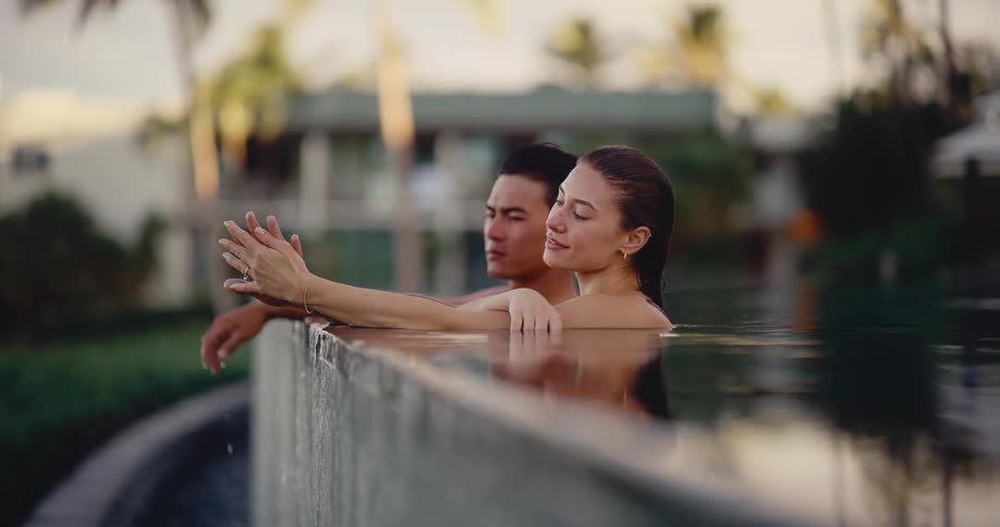 A beautiful young couple holds hands while relaxing in a luxury resort infinity pool at sunset