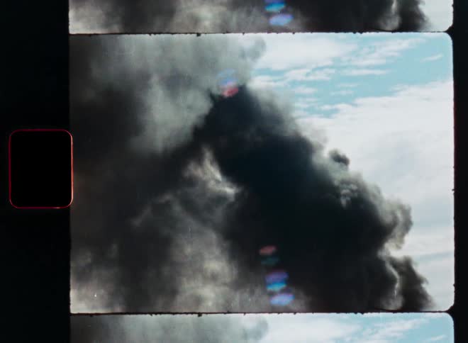 Authentic vintage 8mm film scan shows a thick plume of black smoke billowing into the blue sky, suggesting a fire or explosion