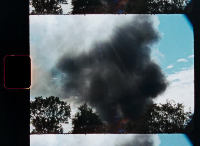 Vintage 8mm film captures thick black smoke rising into the sky from an unseen fire behind a treeline