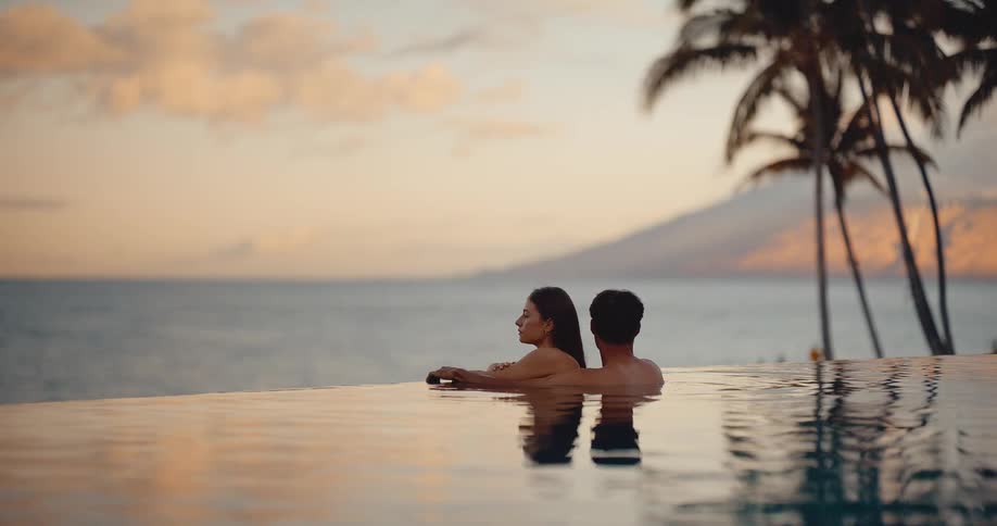 A serene couple relaxes together in a luxurious infinity pool, watching the tranquil sunset over the tropical ocean