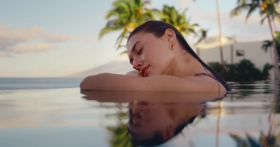 A beautiful young woman relaxes peacefully in a luxurious infinity pool at a tropical resort overlooking the ocean at sunset