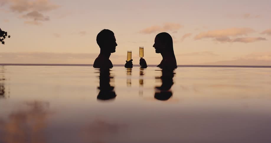 A silhouetted couple enjoys a romantic champagne toast in a luxurious infinity pool as the sun sets over the ocean