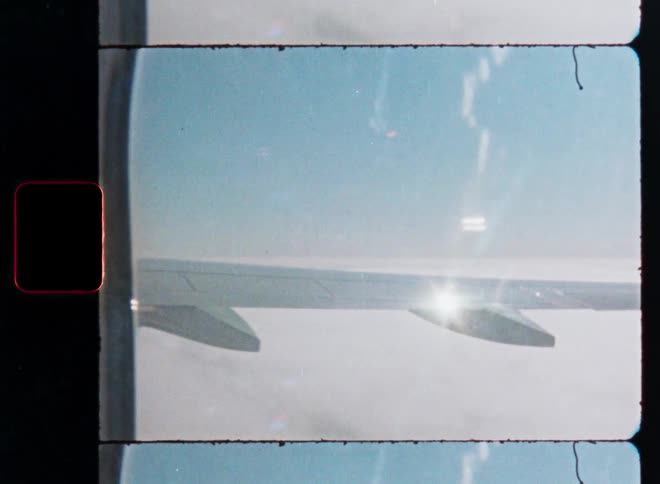 A vintage 8mm film shot from an airplane window shows the wing flying over clouds with a bright sun reflection
