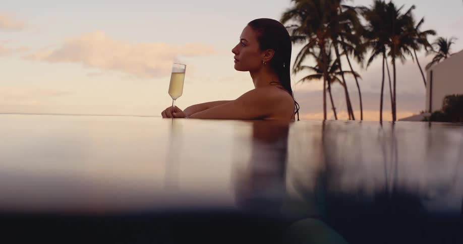 Side profile of an elegant woman relaxing with a glass of champagne in an infinity pool at sunset