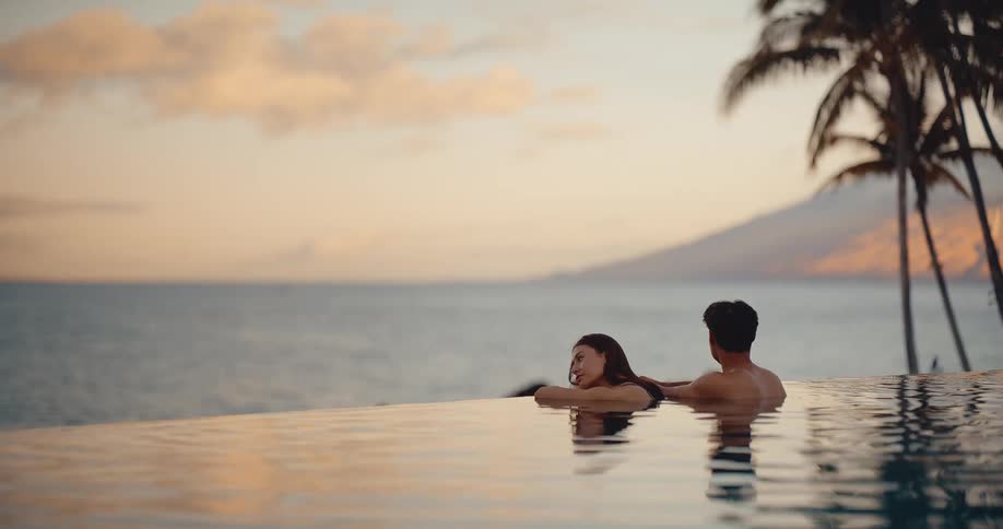 A young couple enjoys a romantic sunset from an infinity pool overlooking the ocean at a tropical luxury resort