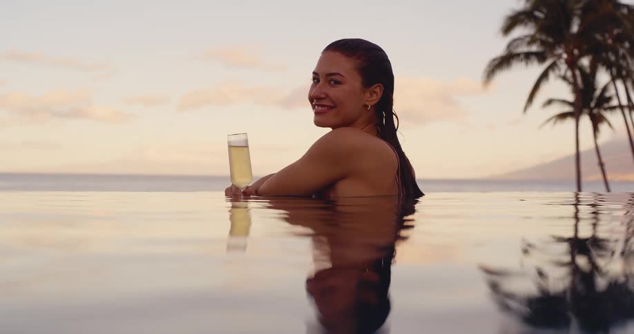A beautiful woman relaxes in an infinity pool with a glass of champagne, watching the tropical sunset over the ocean