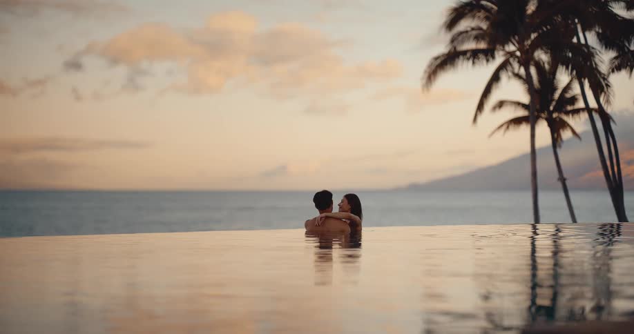 An attractive couple holds each other in a tropical resort infinity pool as they watch the beautiful sunset over the ocean