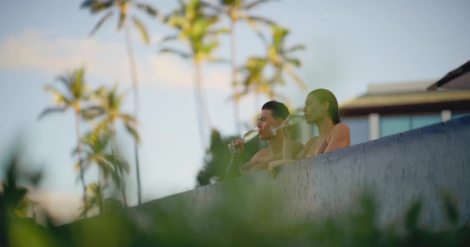 A couple simultaneously sips champagne in slow motion while relaxing in a luxurious tropical resort swimming pool
