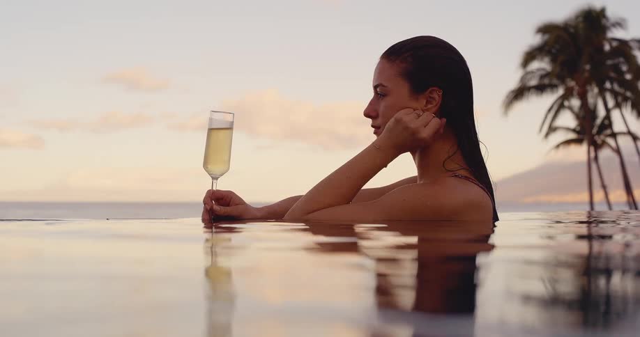 A beautiful woman in an infinity pool holds a glass of champagne and enjoys the tropical ocean sunset in slow motion