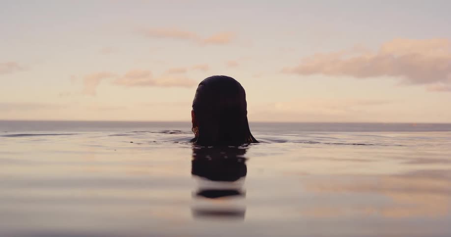 A woman rises gracefully from the water and relaxes in a tranquil infinity pool while watching the beautiful pastel sunset over the ocean