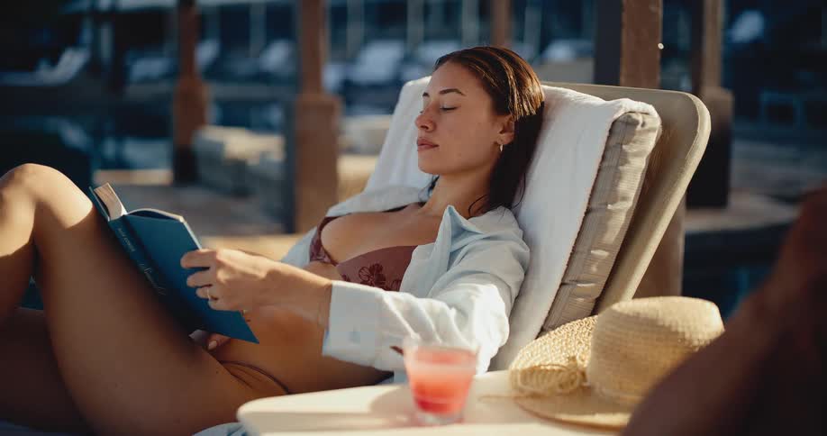 A beautiful woman in a bikini leisurely reads a book on a sun lounger by the pool during her vacation
