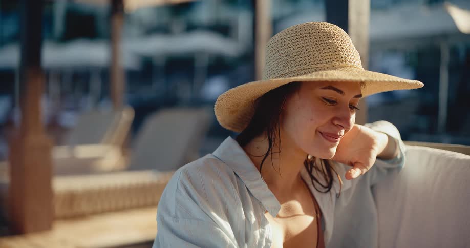 A happy young woman in a straw hat relaxes by the pool with a refreshing cocktail at sunset