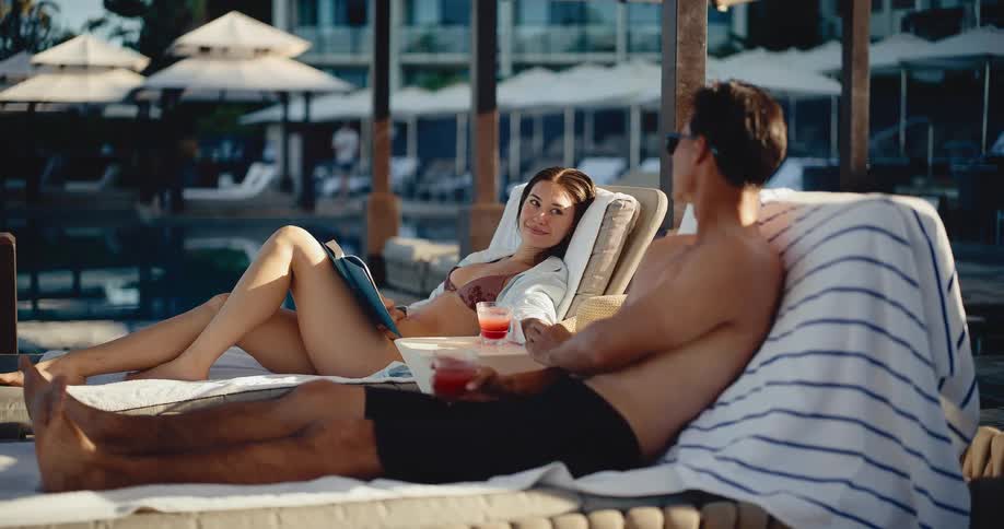 A young couple enjoys a relaxing conversation while lounging by the pool with cocktails at a luxurious resort