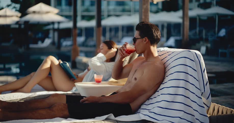 Man and woman relax on lounge chairs by the pool at a luxury resort while enjoying drinks and reading