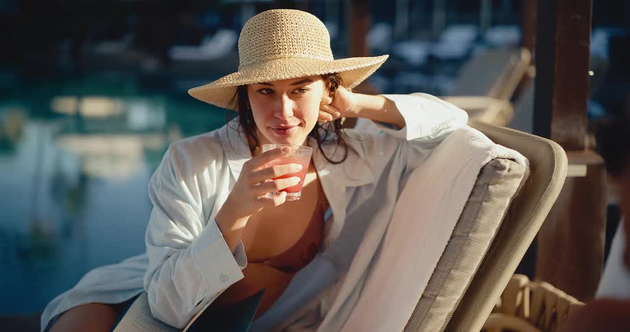 A beautiful woman in a hat relaxes by the pool on vacation, reading a book and sipping a cocktail in slow motion