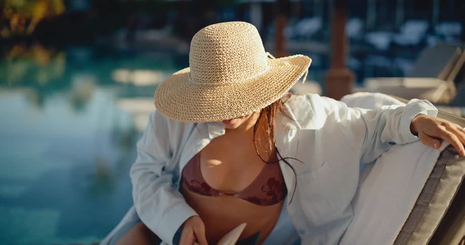 A woman in a bikini and straw hat smiles while relaxing on a lounge chair by a swimming pool