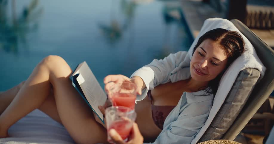 A happy woman in a bikini relaxes on a sun lounger, reading a book, and cheers with a refreshing drink by the pool in slow motion