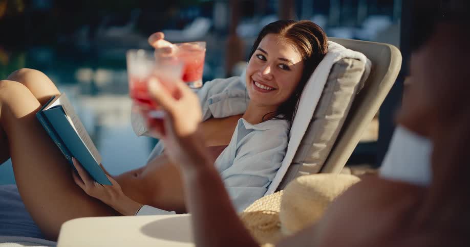 A happy woman reading a book on a lounge chair smiles and toasts her friend with a pink cocktail while relaxing by a luxury resort pool
