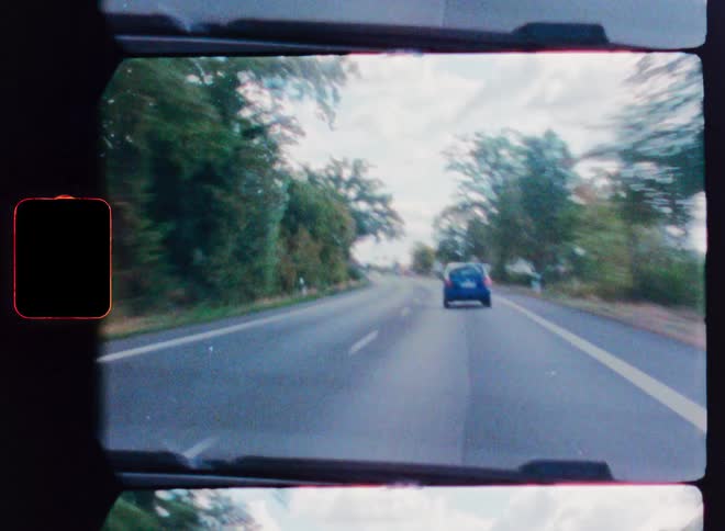 A vintage point-of-view shot from a car driving on a scenic highway, following another vehicle into a suburban area