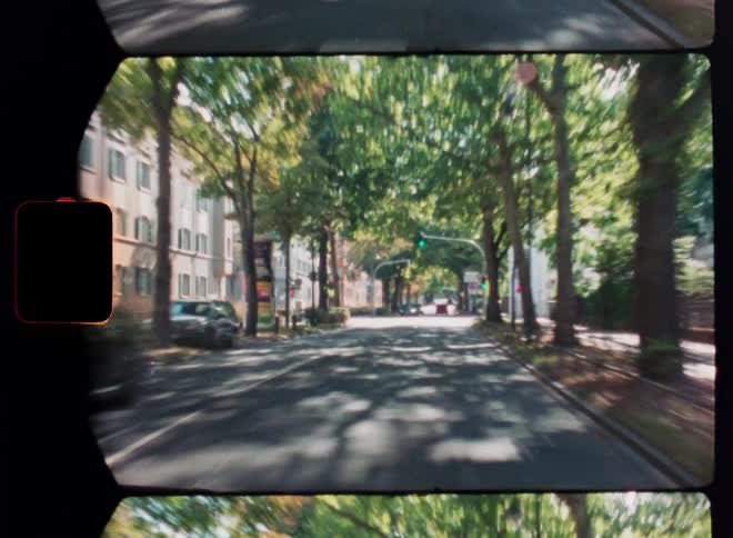 A vintage film-style point-of-view shot of driving down a sunlit, tree-lined street in a European city