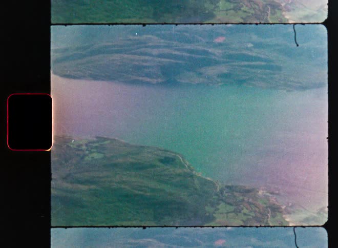 A vintage 8mm film scan shows a mirrored, aerial view of a tranquil lake surrounded by lush green hills