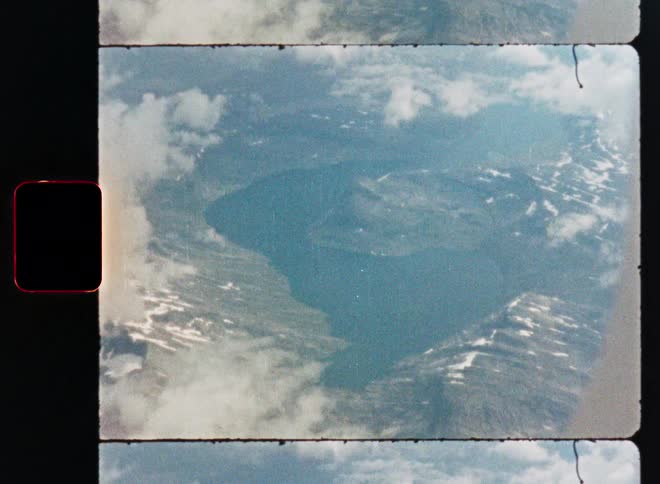A vintage 8mm film scan captures a high-altitude aerial view of a vast crater lake nestled among snowy mountains and clouds