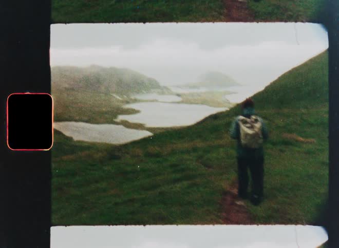 Vintage film of a person with a backpack hiking along a grassy mountain path overlooking a misty valley with lakes