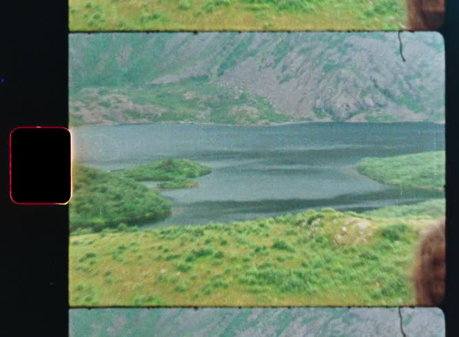 A woman with a backpack looks out at a vast, green mountain landscape with a lake in this vintage, film-style footage
