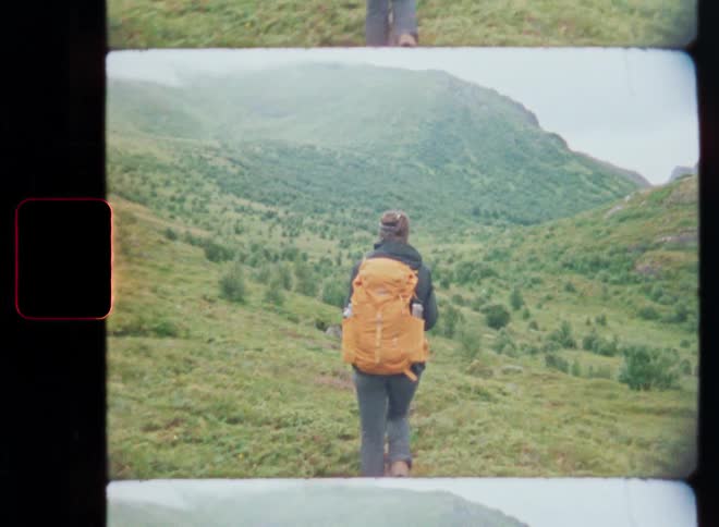 Vintage film captures a hiker with a bright backpack walking along a narrow trail through a verdant mountain valley