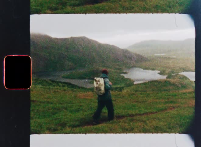 A vintage Super 8 film captures a lone hiker with a backpack exploring a vast, green mountain landscape on an overcast day