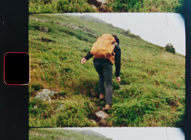 A vintage film scan shows a hiker with a large orange backpack climbing up a steep grassy mountain trail on an overcast day