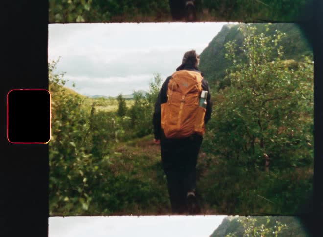 A person with a bright orange backpack hikes through a lush green valley in this vintage-style footage