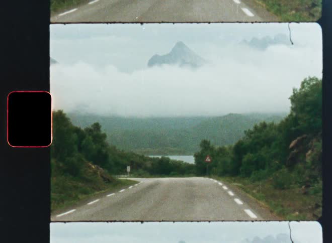 A vintage 8mm film scan captures a scenic road winding towards majestic mountains shrouded in thick fog